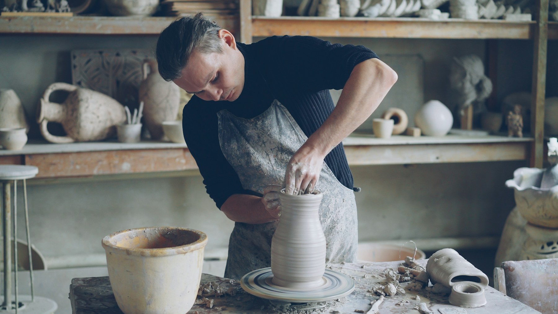 A potter in an apron writing in a notebook at the bench of his studio.
